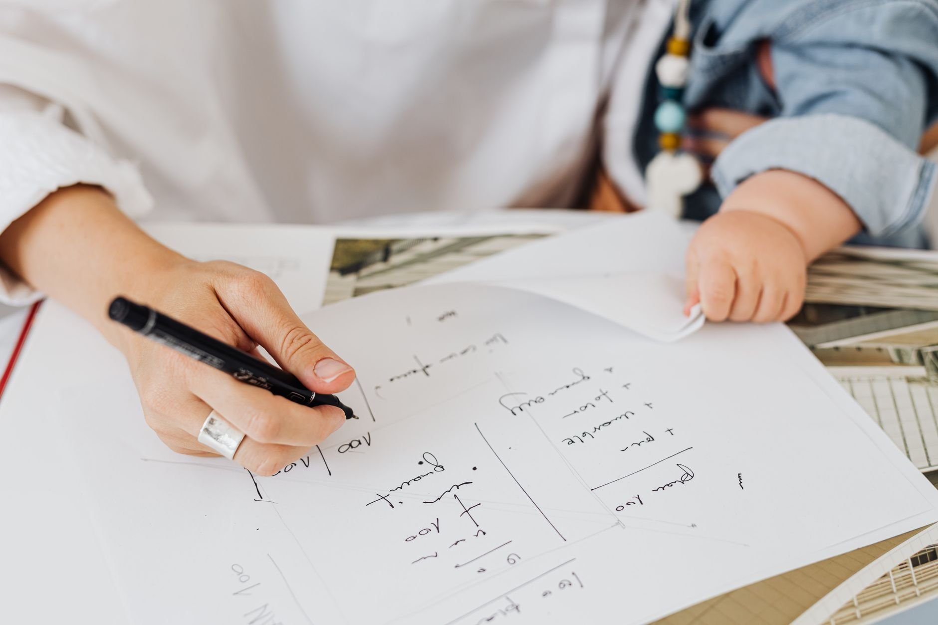 A mother working on papers at home with her baby nearby, in an organized home office setting.