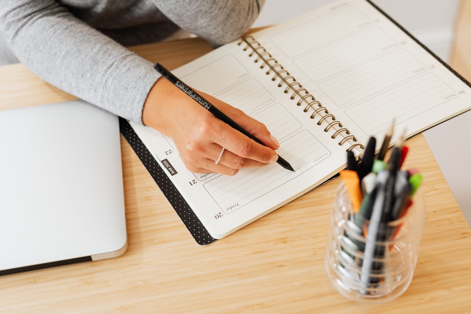 Close-up of a hand writing in a planner on a wooden desk with a laptop nearby.
